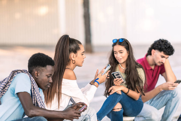 Ryhmä monirotuisia opiskelijoita käyttää matkapuhelinta istuessaan ulkona - Valokuva, kuva