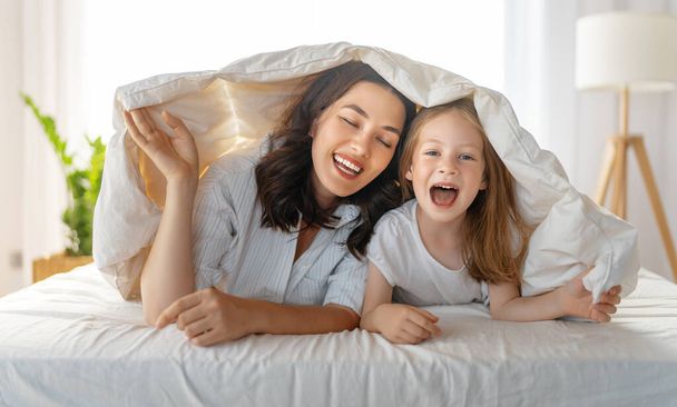 A nice girl and her mother enjoying sunny morning. Good time at home. Child waking up from sleep. Family playing on the bed in the bedroom.  - Photo, image
