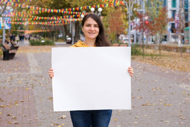 Μια 25χρονη μελαχρινή χαμογελά και κρατά ένα άδειο λευκό σεντόνι για την επιγραφή και το κείμενο - Φωτογραφία, εικόνα