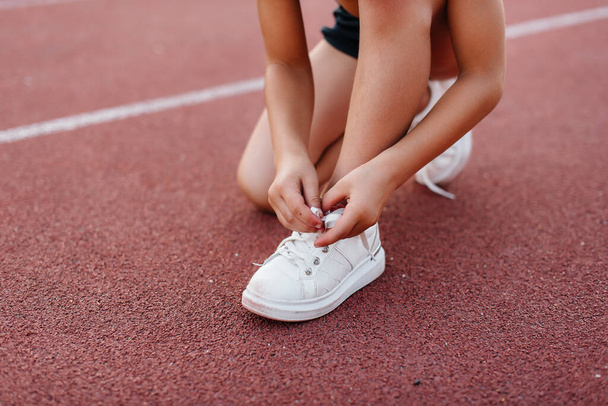 A little girl sat down to tie her shoelaces before running training at the stadium during sunset. Children's sports, and a healthy lifestyle. - Zdjęcie, obraz