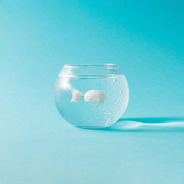 Creative composition with fish bowl and candy wraped in white plastic against blue background. Minimal ecology concept. - Фото, зображення