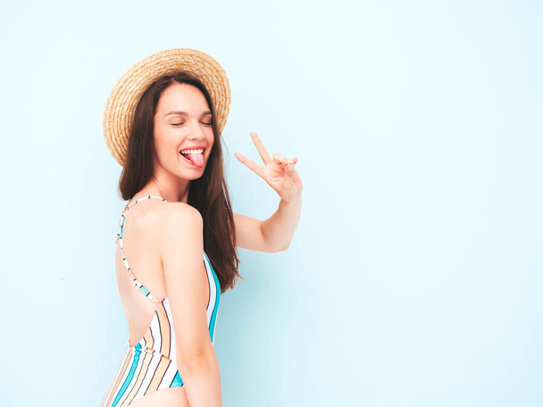 Young beautiful sexy smiling  hipster woman in posing near light blue wall in studio. Trendy model in colorful summer swimwear bathing suit. Positive model shows peace sign.Happy and cheerful in hat - Foto, imagen