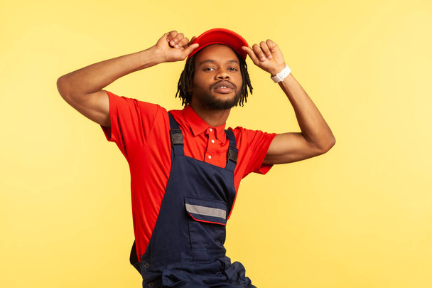 Portrait of stylish worker wearing blue uniform, red T-shirt posing with raised arms, keeps hands on his visor cap, looking directly at camera. Indoor studio shot isolated on yellow background. - Photo, image