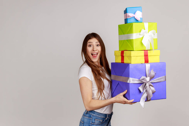 Surprised excited woman holding lot of wrapped boxes and looking with amazement, shocked by mount of bonuses and gifts, wearing white T-shirt. Indoor studio shot isolated on gray background. - Φωτογραφία, εικόνα