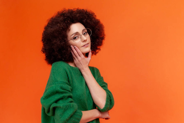 Profile portrait of beautiful woman with Afro hairstyle in green casual style sweater and eyeglasses, looking at camera, touching her face, skin care. Indoor studio shot isolated on orange background. - Photo, image