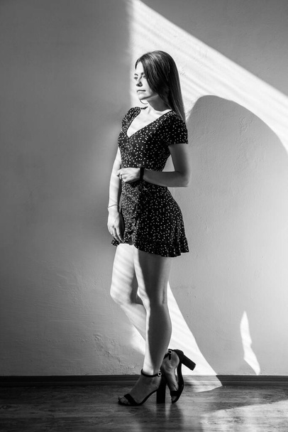 Attractive dark haired female standing looking away with romantic expression, wearing dress and sandals. Black and white photography, indoor studio shot illuminated by sunlight from window. - Fotoğraf, Görsel
