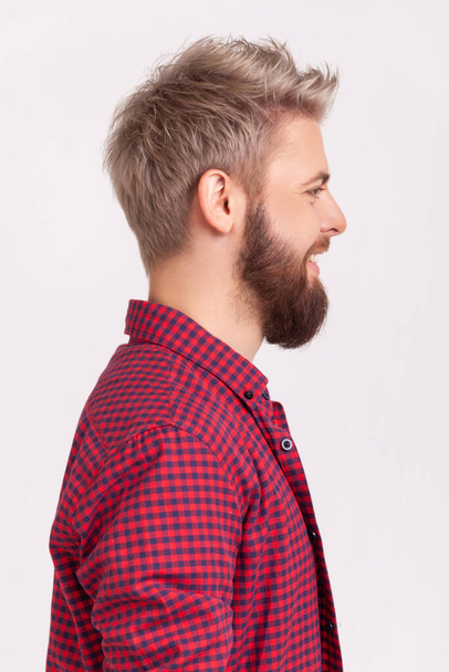 Profile portrait of happy bearded millennial man in red t-shirt sincerely smiling and looking to side, positive mood and optimistic lifestyle. Indoor studio shot isolated on gray background - Valokuva, kuva