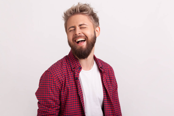Portrait of funny handsome bearded young man in plaid shirt standing with closed eyes and laughing. Indoor studio shot isolated on gray background - Photo, Image