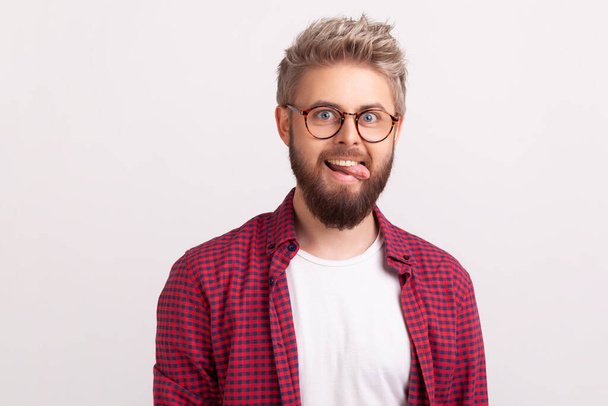 Portrait of funny crazy handsome bearded man in eyeglasses and plaid shirt fooling around and showing tongue, optimism. Indoor studio shot isolated on gray background - Foto, immagini