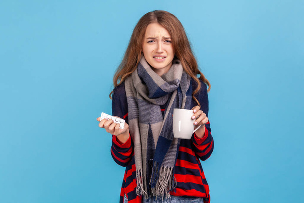 Sick woman wearing striped casual style sweater, wrapped in scarf holding pills and tea cup, screaming, feeling annoyed from virus grippe. Indoor studio shot isolated on blue background. - Foto, Bild