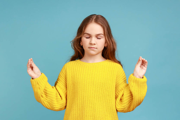 Portrait of little girl holding arms in mudra gesture, feeling calm relaxed with peaceful expression, wearing yellow casual style sweater. Indoor studio shot isolated on blue background. - Fotó, kép