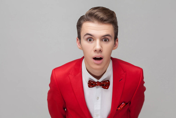 Portrait of handsome gentleman with stylish hairdo in red tuxedo and bow tie standing with big eyes and open mouth, looking at camera in amazement, shock. studio shot isolated on gray background - Fotó, kép