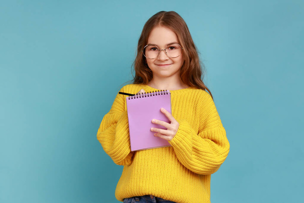 Portrait of positive little girl writing in notebook, looks smiling at camera, child doing homework, wearing yellow casual style sweater. Indoor studio shot isolated on blue background. - Foto, Bild