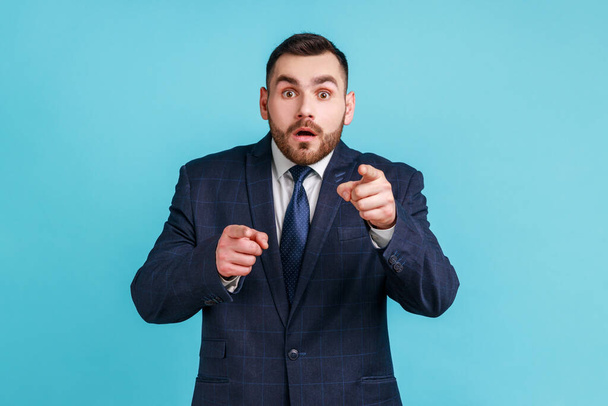 Amazed astonished bearded man wearing official style suit pointing finger to camera, looking with interrogative expression at camera. Indoor studio shot isolated on blue background. - Fotoğraf, Görsel