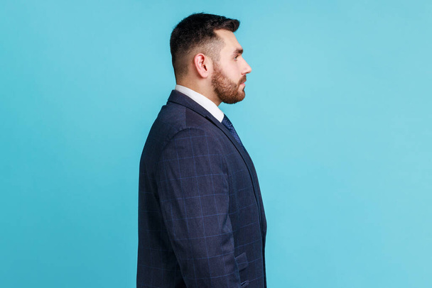 Side view of bearded businessman wearing dark official style suit, having serious facial expression, being upset, has problems at work. Indoor studio shot isolated on blue background. - Photo, Image