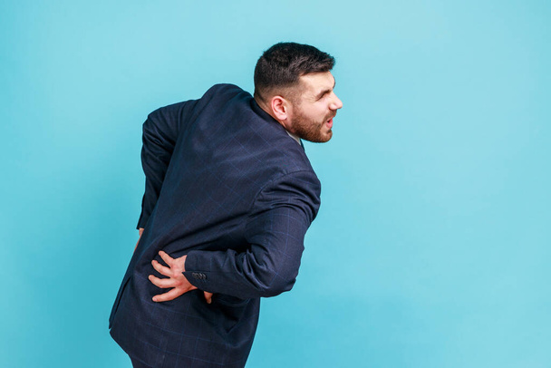 Spine problems. Young adult businessman in official style suit holding his back, having sudden lower back pain, pinched nerve or kidney inflammation. Indoor studio shot isolated on blue background. - Foto, Imagen