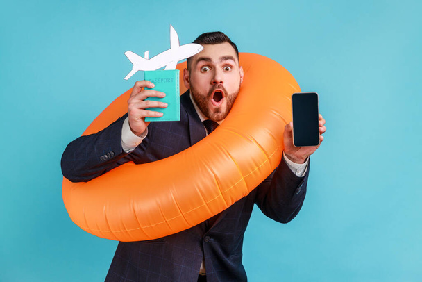 Portrait of young adult ma with beard wearing official style suit holding orange rubber ring, paper plane, passport, planning vacation abroad. Indoor studio shot isolated on blue background. - Photo, image