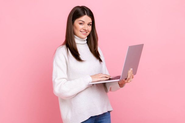 Successful office worker girl looking at camera with happy face, holding laptop, typing on keyboard, wearing white casual style sweater. Indoor studio shot isolated on pink background. - Fotó, kép