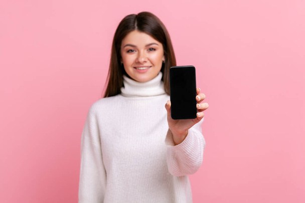 Smiling brunette female showing smartphone with blank screen for advertisement or promotional text, wearing white casual style sweater. Indoor studio shot isolated on pink background. - Fotografie, Obrázek