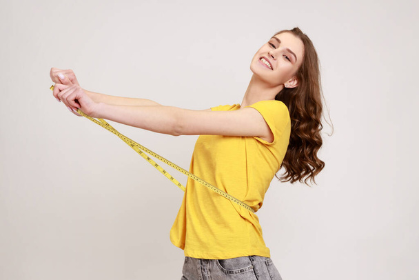 Profile portrait happy woman of toothy smile with toothy smile in yellow T-shirt measuring her waist with tape measure, satisfied with result. Indoor studio shot isolated on gray background. - 写真・画像