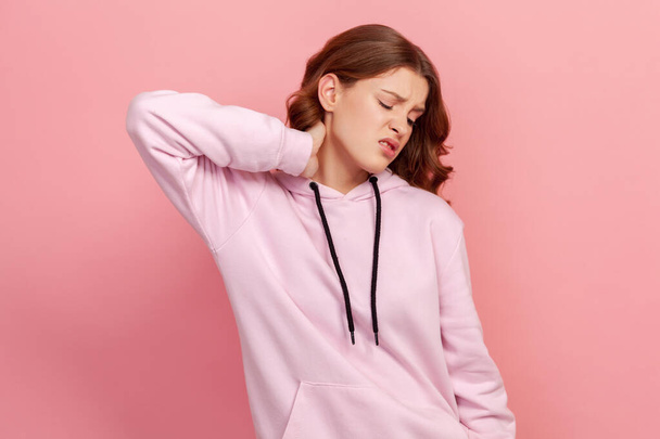 Portrait of upset brunette teen girl in hoodie touching neck feeling pain and numbness, worried about muscle tension, osteochondrosis. Indoor studio shot isolated on pink background - Foto, Bild