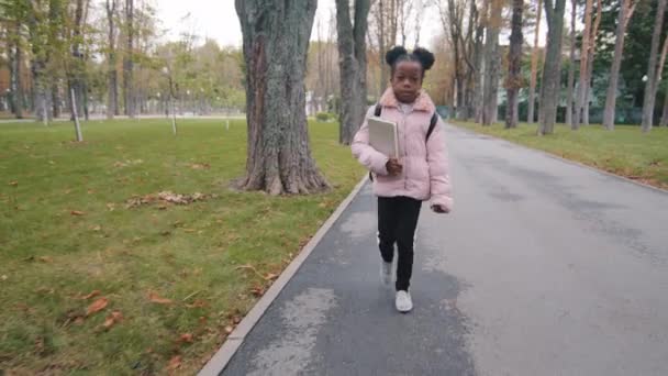 Маленька афро-американська дівчинка-афроамериканка, учениця-афроамериканка з рюкзаком, яка ходить по вулиці в парку, йде на урок назад до школи. - Кадри, відео
