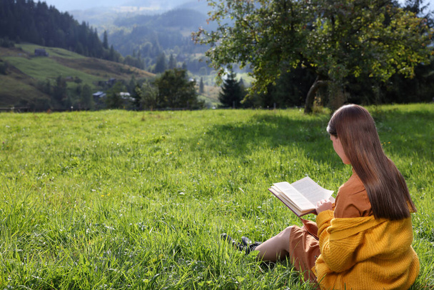 Young woman reading book on green meadow in mountains, back view - Zdjęcie, obraz