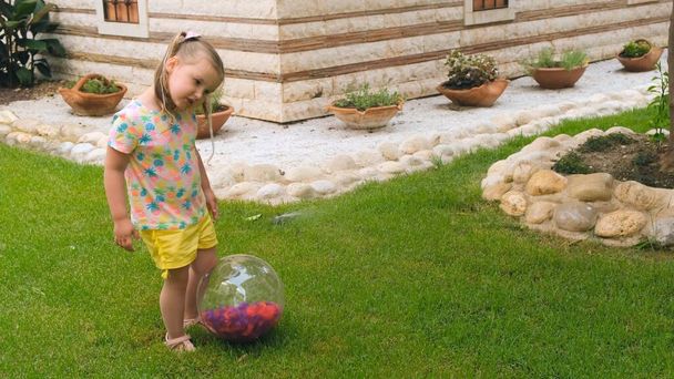 смешная милая маленькая девочка, 3 года, с двумя хвостиками на голове, в разноцветной футболке и желтых шортах, играет с разноцветным мячом на зеленом лугу в саду ее - Фото, изображение