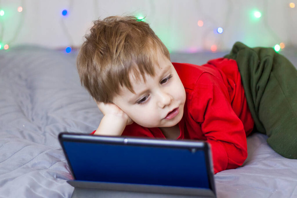 A cute boy is sitting on the bed and looking at a tablet. Funny smiling child. The concept of gadgets and children. - 写真・画像