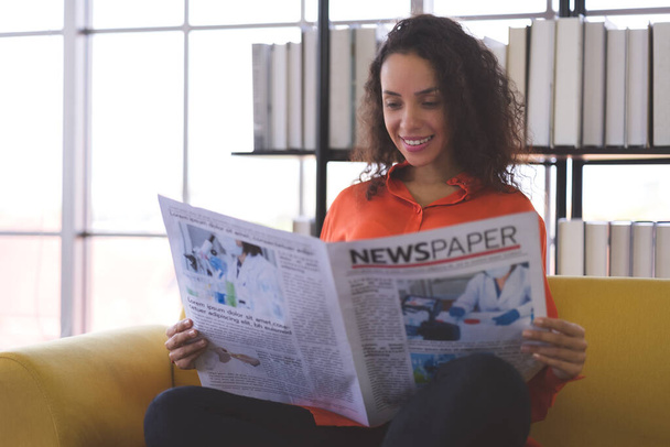 Mooie jonge zwarte vrouw draagt oranje hemd glimlachend en leest thuis nieuwsberichten op de bank. Zakelijke vrouw en freelance werk van huis in de verspreiding van coronavirus covid-19. - Foto, afbeelding