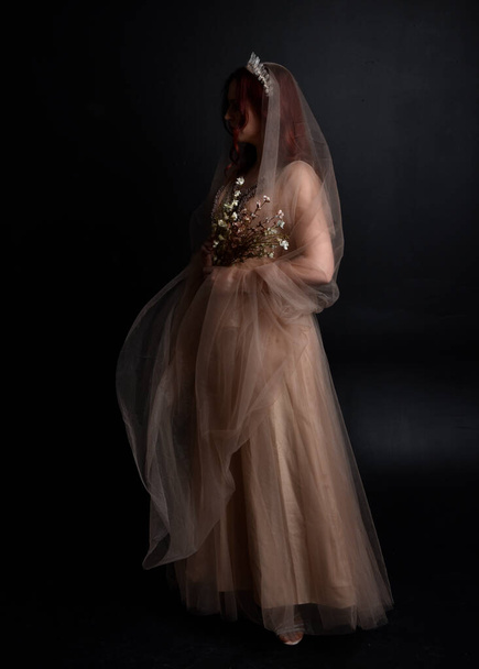 Plně dlouhý portrét rudovlasé dívky v krémových fantasy šatech jako kostým víly bohyně. stojící póza s elegantním gesturálním pohybem pozpátku kamery, izolované na tmavém pozadí studia. - Fotografie, Obrázek