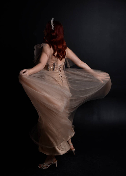 Pełna długość portret rudej dziewczyny w kremowej sukni fantazji jak kostium bogini bajki. pozycja stojąca z eleganckim ruchem gestykulacyjnym tyłem do kamery, odizolowana na ciemnym tle studia. - Zdjęcie, obraz