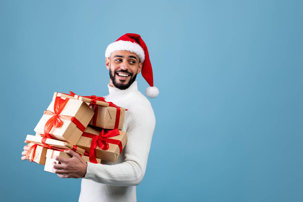 Feiertage und Feiertage. Glücklicher arabischer Kerl mit Weihnachtsmütze, Geschenke in der Hand und Blick auf freien Raum, blauer Hintergrund - Foto, Bild