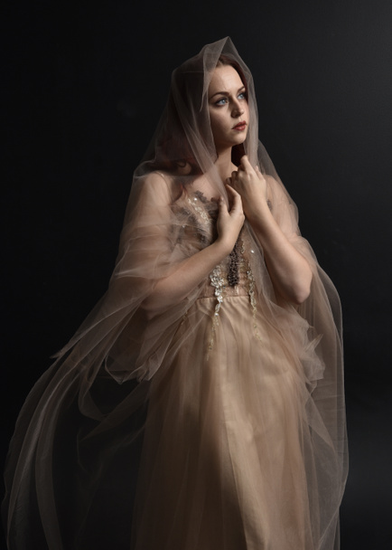 Full length  portrait of red haired  girl wearing a creamy fantasy gown like a fairy goddess costume.  standing  pose with elegant gestural movement , isolated on dark studio background. - 写真・画像