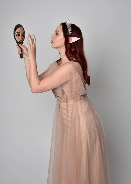 Full length  portrait of red haired  girl wearing a creamy fantasy gown and crystal crown, like a fairy goddess costume.  standing  pose with elegant gestural arm movement holding a golden mirror , isolated on light studio background. - Photo, Image
