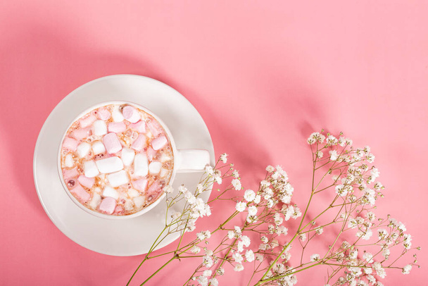 Cup of cocoa with marshmallows and white flowers on a pink background, top view, free space for text - Fotó, kép