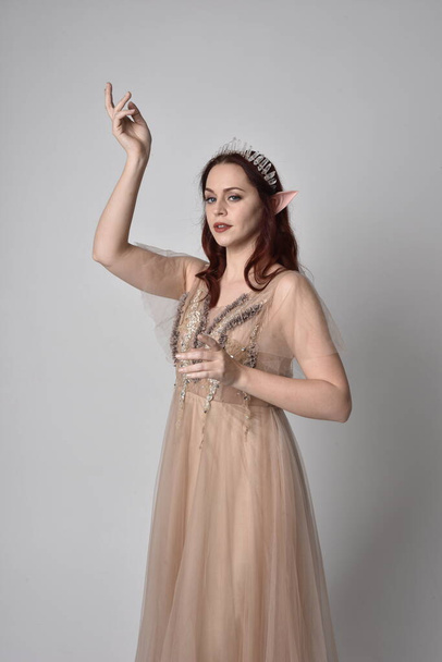 Full length  portrait of red haired  girl wearing a creamy fantasy gown and crystal crown, like a fairy goddess costume.  standing  pose with elegant gestural hands, isolated on light studio back - Φωτογραφία, εικόνα