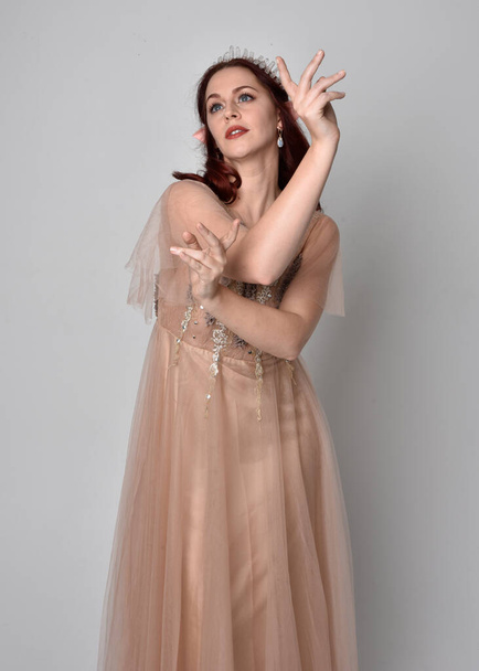 Full length  portrait of red haired  girl wearing a creamy fantasy gown and crystal crown, like a fairy goddess costume.  standing  pose with elegant gestural hands, isolated on light studio back - Photo, Image