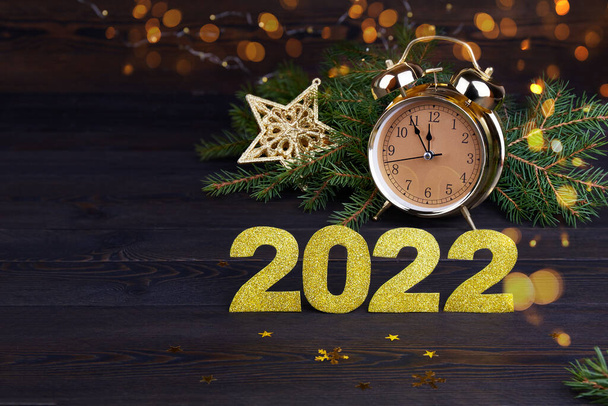 Happy New Year 2022 Greeting Card. Fir branches, golden numbers 2022 , bokeh, sparkling lights, alarm clock on dark wooden background.  - 写真・画像