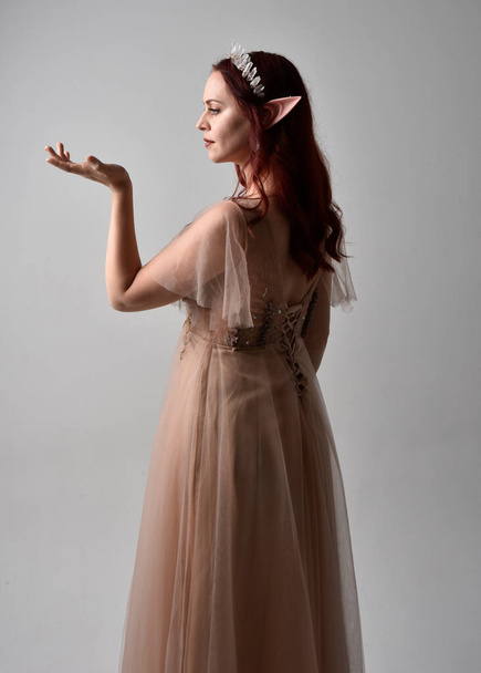 Retrato de comprimento total da menina de cabelos vermelhos vestindo um vestido de fantasia cremoso e coroa de cristal, como um traje de fada deusa. postura de pé com mãos gestuais elegantes, isolado no estúdio de luz de volta - Foto, Imagem