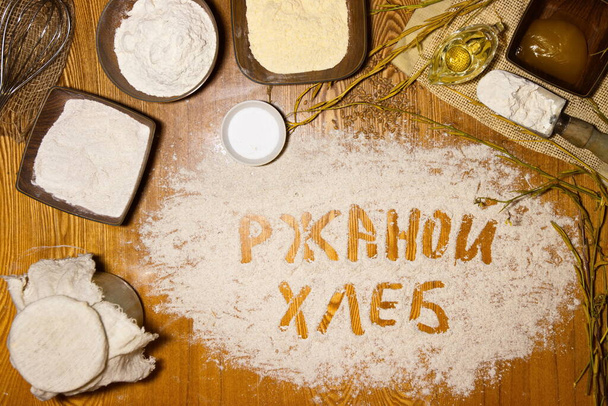 Σπιτικό ψωμί. Επίπεδη. Αλεύρι είναι διάσπαρτα στο τραπέζι στο οποίο "ψωμί σίκαλης" είναι γραμμένο στα ρωσικά. Στο τραπέζι υπάρχουν συστατικά για την παρασκευή ψωμί προζύμης χωρίς μαγιά σίκαλης: αλεύρι (σίκαλη, σιτάρι, καλαμπόκι) προζύμη, αλάτι, μέλι, λάδι. - Φωτογραφία, εικόνα