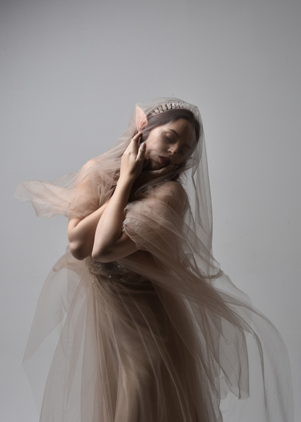 portrait of red haired  girl wearing a creamy fantasy gown and flowing veil fabric, like a fairy goddess costume.  standing  pose with elegant gestural hands, isolated on light studio background. - Foto, Bild