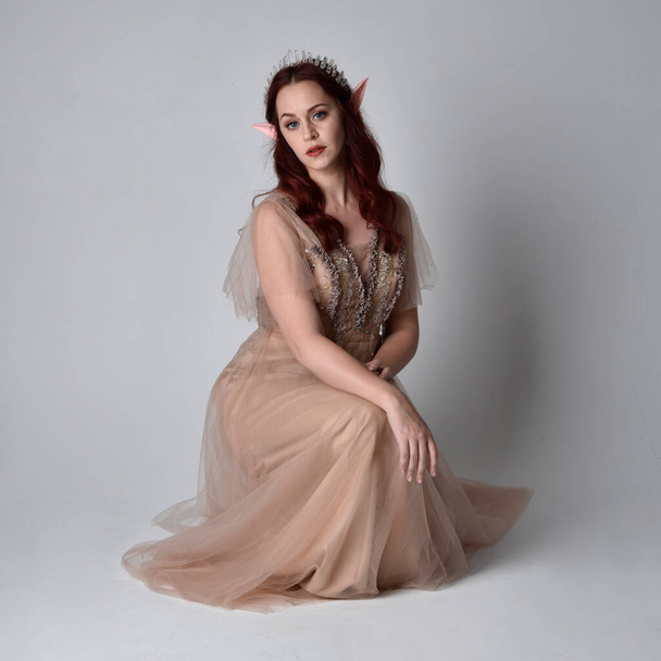 Full length  portrait of red haired  girl wearing a creamy fantasy gown and crystal crown, like a fairy goddess costume.  sitting pose with elegant gestural hands, isolated on light studio background. - Фото, изображение