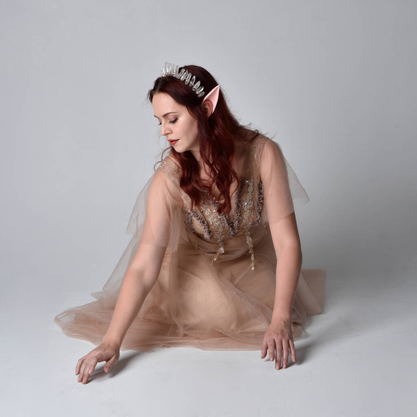 Full length  portrait of red haired  girl wearing a creamy fantasy gown and crystal crown, like a fairy goddess costume.  sitting pose with elegant gestural hands, isolated on light studio background. - Foto, Imagen