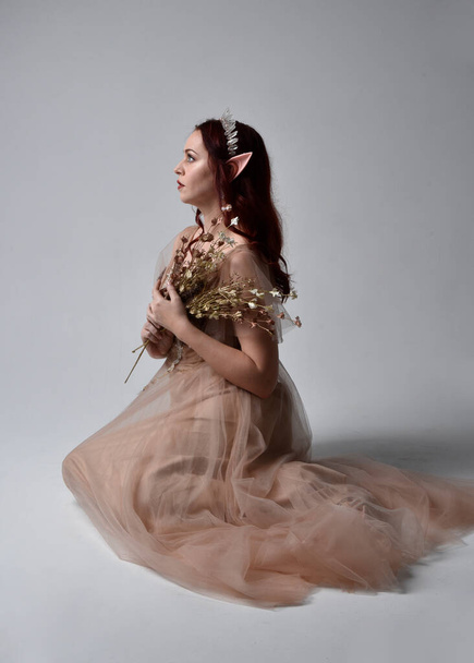 Full length  portrait of red haired  girl wearing a creamy fantasy gown and crystal crown, like a fairy goddess costume.  sitting pose with elegant gestural hands, isolated on light studio background. - Foto, Imagen