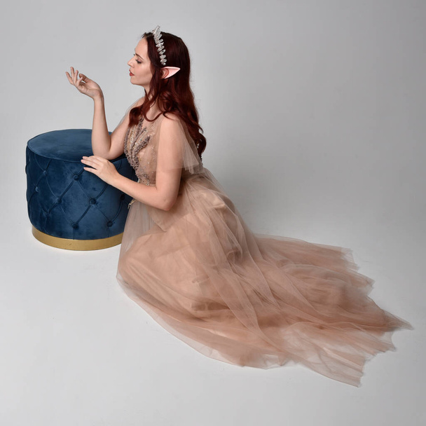 Портрет рудої волохатої дівчини у кремовому фентезійному сукні та кришталевій короні, як костюм феї. сидяча поза з елегантними жестовими руками, ізольована на світлому фоні студії
. - Фото, зображення