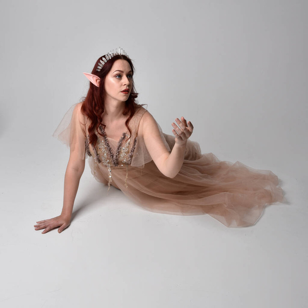 Full length  portrait of red haired  girl wearing a creamy fantasy gown and crystal crown, like a fairy goddess costume.  sitting pose with elegant gestural hands, isolated on light studio background. - 写真・画像