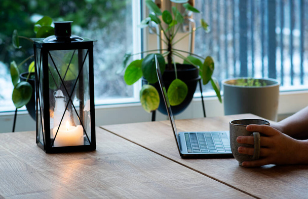 Человек работает дома в зимний день, пользуется ноутбуком и пьет кофе. Свечной фонарь стоит на деревянном столе. - Фото, изображение