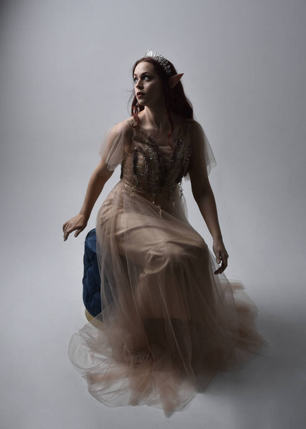 Full length  portrait of red haired  girl wearing a creamy fantasy gown and crystal crown, like a fairy goddess costume.  sitting pose with elegant gestural hands, isolated on light studio background. - Foto, imagen