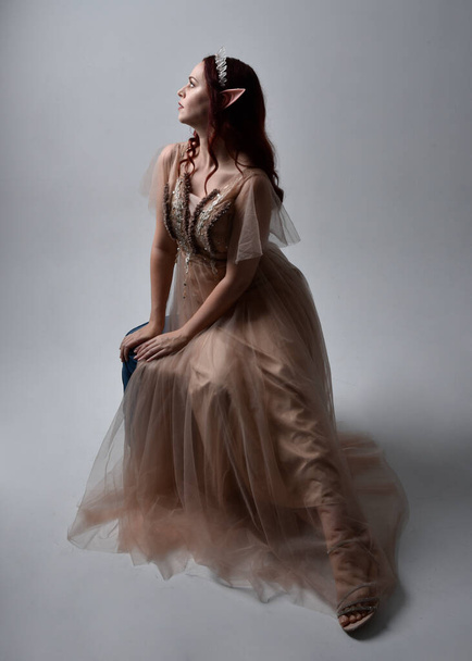 Portrait complet de fille aux cheveux roux portant une robe fantaisie crémeuse et une couronne de cristal, comme un costume de déesse fée. pose assise avec des mains gestuelles élégantes, isolée sur fond de studio lumineux. - Photo, image